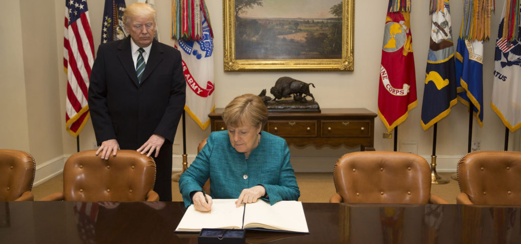 Donald Trump stands behind Angela Merkel as she writes in the White House guest book. Neither of them looks happy about it.