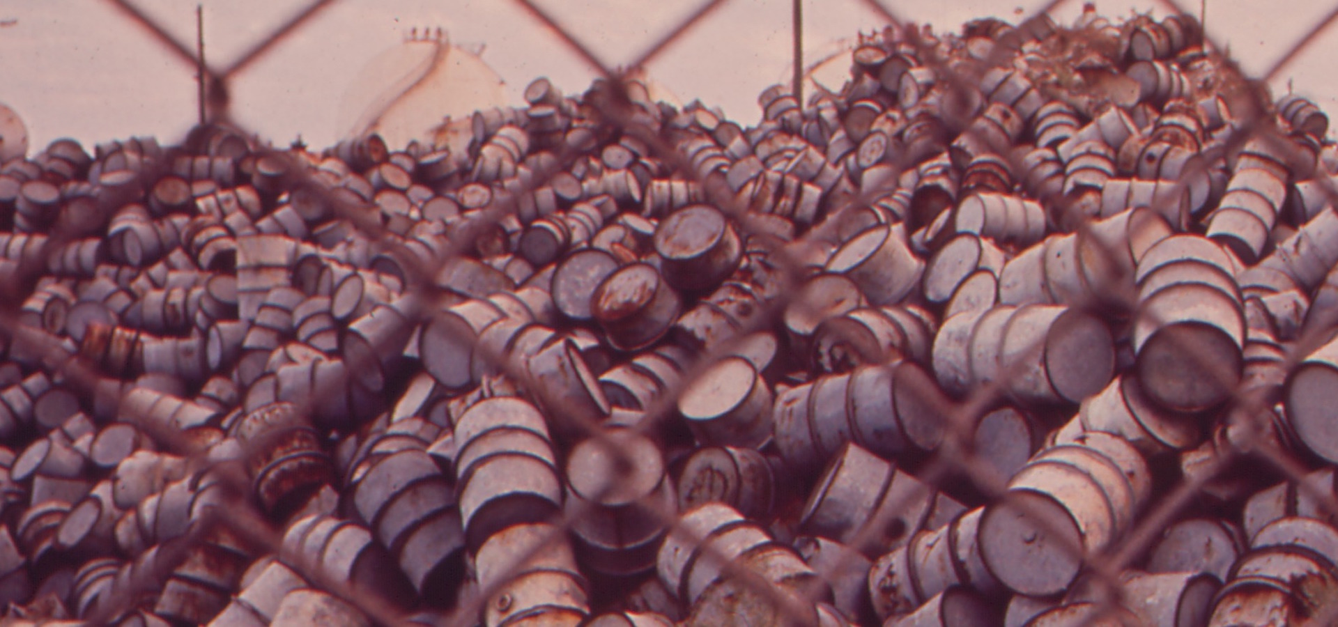 a mountain of damaged oil drums seen through a chain link fence