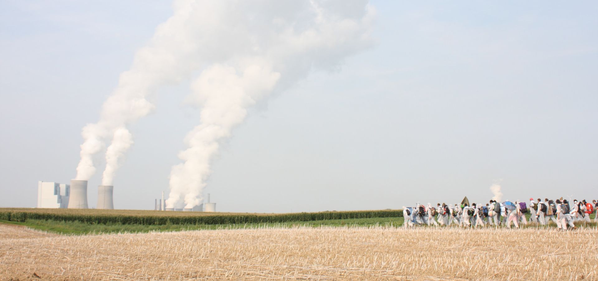 group of activists in white jumpsuits walking through a field towards coal towers