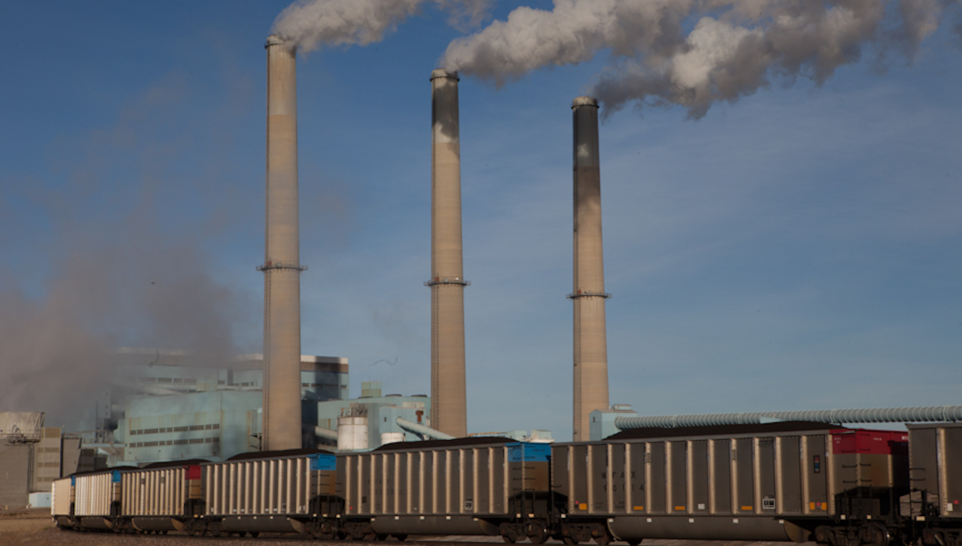 three smokestacks emit clouds with a train carrying coal underneath