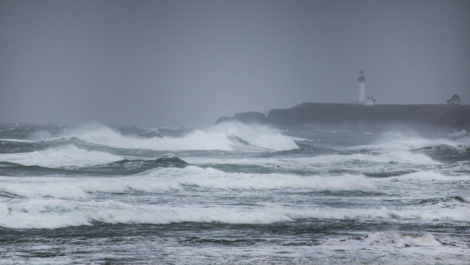 huge waves and gray sky with a lighthouse visible in the background at Yaquina 